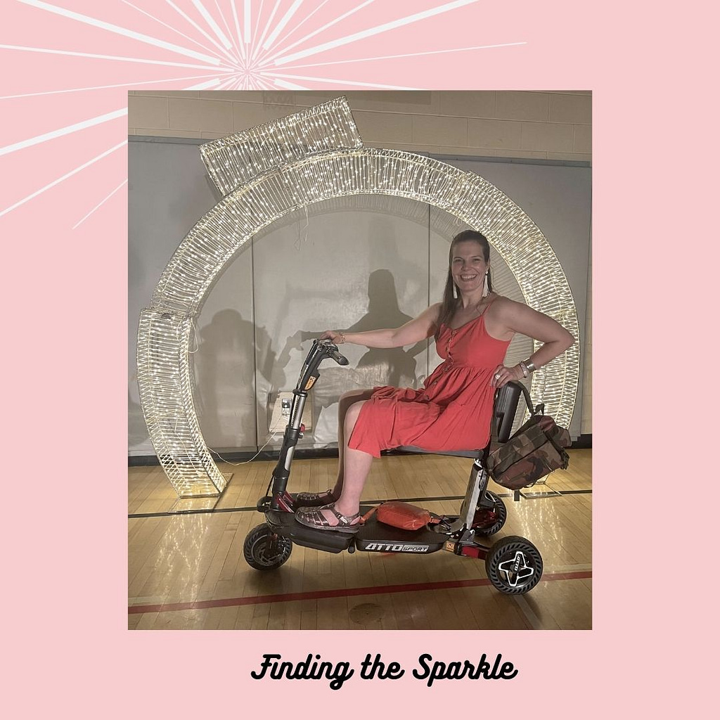 ME sitting on my Atto Mobility circle in front of a twinkling semi circle. I am a white female with long brow hair. I am wearing a red farm style dress with spaghetti straps.