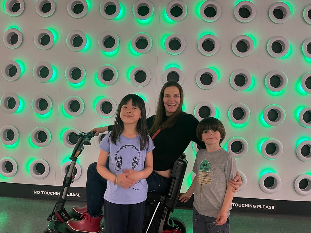 ME on my Atto Scooter at the Vancouver Science centre. I am flanked by my 6 year old son to the right and his friend a pretty 6 year old girl to the right. We are standing infant of a wall of illuminated green circles.