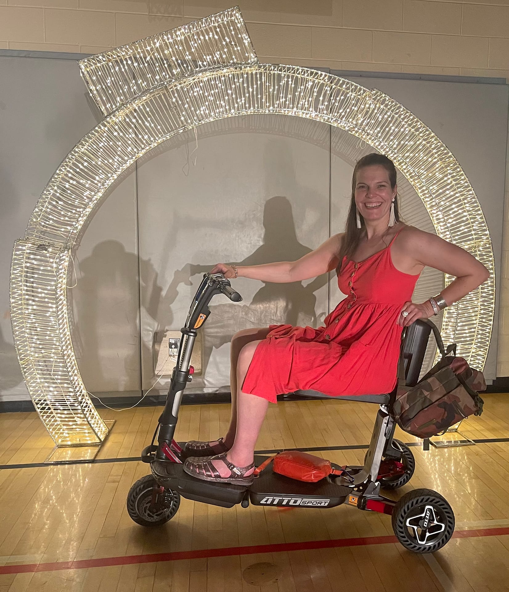 Me wearing a red dress and jelly shoes. I am a caucasian woman with long brown hair sitting on my Atto Mobility Scooter. I have stopped in front of a large sparkly semi circle. I have a big smile on my face.